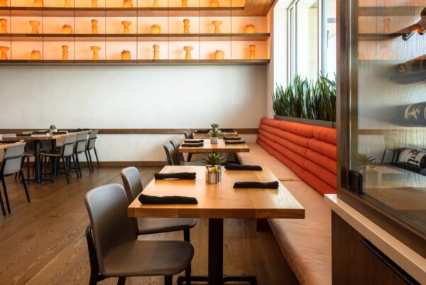 A modern dining area with light wood tables and grey chairs, featuring a long orange cushioned bench against the wall, and shelves with illuminated orange ceramic vases.