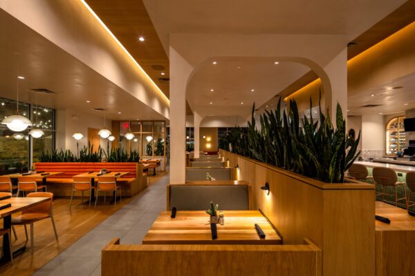 Modern restaurant interior with warm lighting, featuring booth seating on the right and a row of tables with orange cushioned seating on the left, separated by tall greenery.