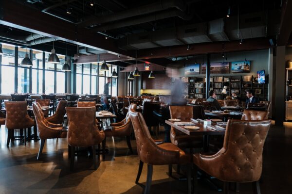 A spacious dining area with leather chairs and wooden tables under industrial pendant lighting, with large windows providing natural light.