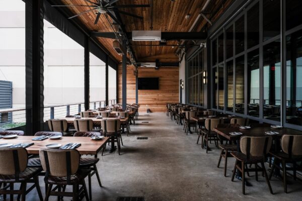 A beautifully designed dining space furnished by South Loft, featuring wooden tables and comfortable upholstered chairs under a warm wooden ceiling. The outdoor patio area is partially enclosed with large glass windows and industrial accents, creating a cozy yet modern atmosphere.