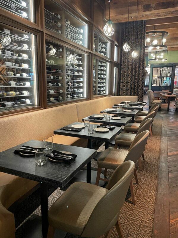 A sophisticated dining area furnished by South Loft featuring a row of modern black tables paired with elegant upholstered chairs. The backdrop includes a well-lit wine display encased in glass, showcasing an extensive wine collection, complemented by warm lighting and luxurious decor.